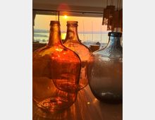 Große Glasflaschen in Bernstein- und Blautönen stehen im Vordergrund auf einem Tisch, dahinter ein Innenraum mit Hängeleuchten und großer Fensterfront. Durch die Fenster ist ein Sonnenuntergang über einem Hafen mit Segelmasten und Wasser zu sehen.