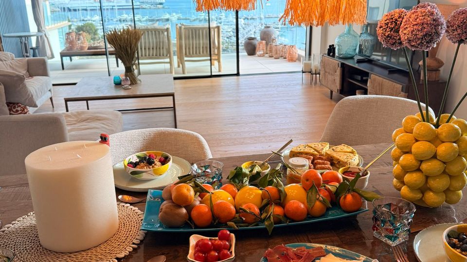 Offener Wohn- und Essbereich mit großem Holztisch und gedecktem Frühstück, darüber mehrere große, fransenartige Hängelampen. Im Hintergrund führen bodentiefe Glasschiebetüren auf eine Terrasse mit Blick über einen Hafen und das Meer.