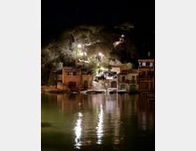 Vista nocturna d’un petit port o cala amb cases de façana clara i teulades de teula a tocar de l’aigua, amb portes i arcs a nivell del moll. Al fons hi ha una escala il·luminada amb fanals que puja entre arbres, i els llums es reflecteixen sobre la superfície tranquil·la de l’aigua.