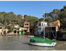 Petita cala amb cases tradicionals de façanes clares i teulades de teula, amb portes verdes a nivell de l’aigua i un bosc de pins al darrere. En primer terme hi ha un veler amarrat amb el casc blanc i una lona verda, sobre aigua tranquil·la.