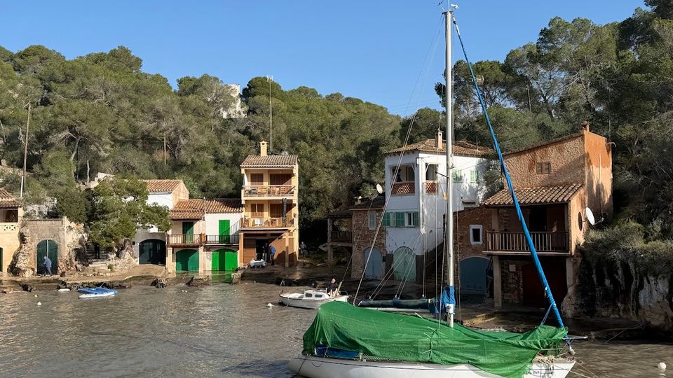 Petita cala amb cases tradicionals de façanes clares i teulades de teula, amb portes verdes a nivell de l’aigua i un bosc de pins al darrere. En primer terme hi ha un veler amarrat amb el casc blanc i una lona verda, sobre aigua tranquil·la.