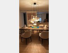 Moderner Essbereich mit langem Holztisch, gepolsterten Stühlen und einer zentral hängenden, mehrteiligen Pendelleuchte mit warmem Licht. Im Hintergrund sind eine offene Küche mit Tresen, dekorative Vasen und ein großformatiges Wandbild zu sehen.