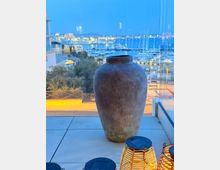 Blick von einer verglasten Terrasse auf einen beleuchteten Yachthafen mit vielen Masten und Booten in der Dämmerung. Im Vordergrund stehen eine große Tonvase auf Fliesen sowie kleine Laternen am Boden.