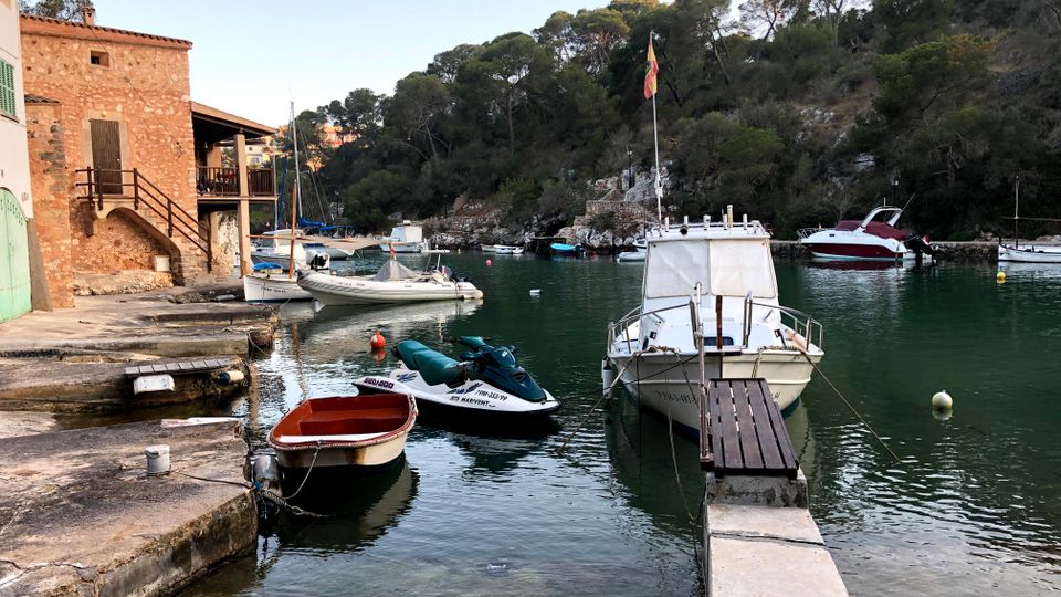 Petit port en una cala d’aigües tranquil·les, amb diverses embarcacions amarrades a un moll de pedra i una passarel·la estreta cap a un vaixell blanc. A l’esquerra hi ha edificis de pedra vora l’aigua i al fons un vessant boscós amb roques i pins que envolta la badia.