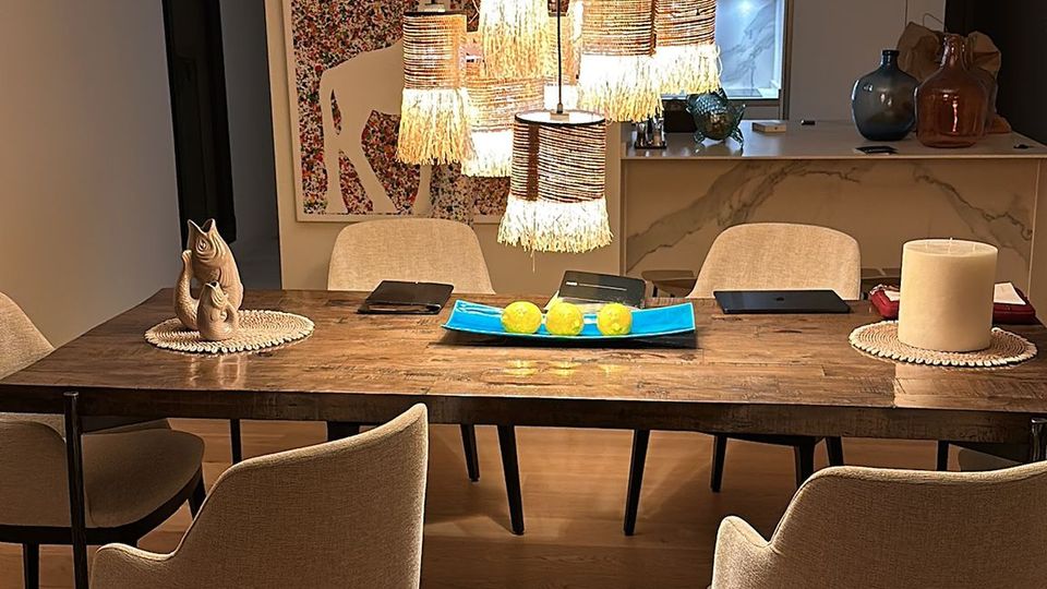 Moderner Essbereich mit langem Holztisch, gepolsterten Stühlen und einer zentral hängenden, mehrteiligen Pendelleuchte mit warmem Licht. Im Hintergrund sind eine offene Küche mit Tresen, dekorative Vasen und ein großformatiges Wandbild zu sehen.