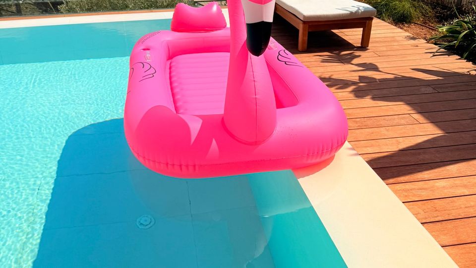 Infinity-Pool mit türkisfarbenem Wasser neben einer Holzterrasse mit Liege; im Wasser schwimmt ein großer pinker Flamingo-Schwimmreifen. Dahinter Blick über eine Marina mit Segelbooten und Yachten auf das Meer und eine Küstenstadt unter blauem Himmel.