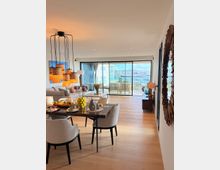 Offener Wohn- und Essbereich in einer modernen Wohnung mit hellem Holzboden, Esstisch mit Polsterstühlen und Sofaecke unter mehreren hängenden Lampenschirmen. Große Schiebetüren führen auf einen Balkon; durch die Glasfront ist ein Blick auf eine Marina und das Wasser zu sehen.