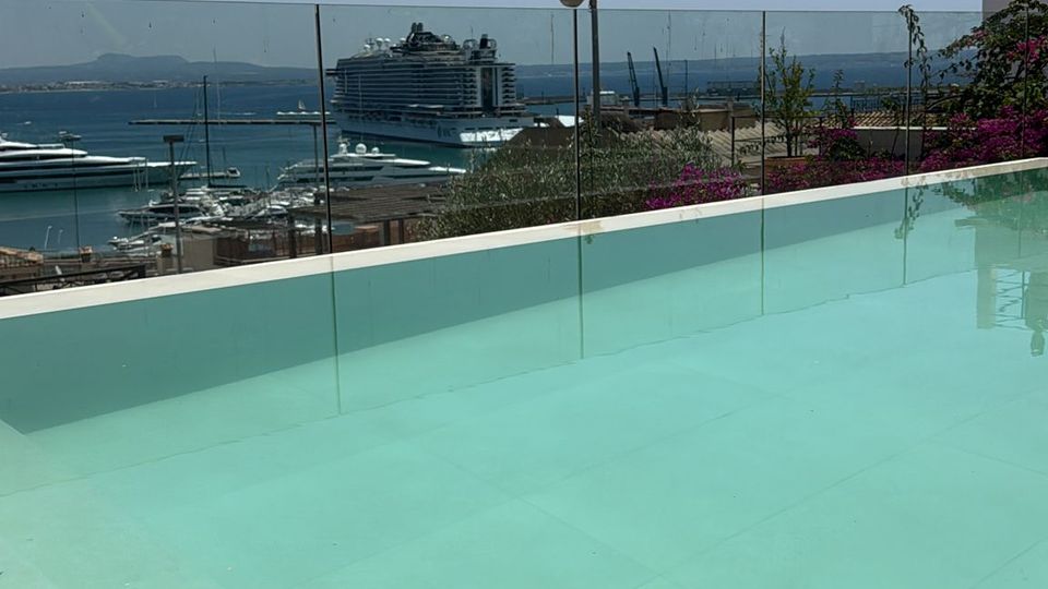 Infinity-Pool auf einer Dachterrasse mit türkisfarbenem Wasser und Glasgeländer, im Hintergrund ein Hafen mit Kreuzfahrtschiff und Yachten. Dahinter erstrecken sich Meer und Küstenhügel unter klarem Himmel.