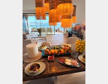 Offener Wohn- und Essbereich mit großem Holztisch und gedecktem Frühstück, darüber mehrere große, fransenartige Hängelampen. Im Hintergrund führen bodentiefe Glasschiebetüren auf eine Terrasse mit Blick über einen Hafen und das Meer.