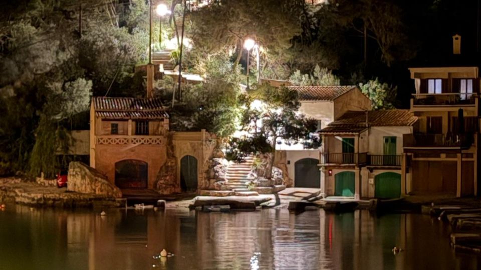 Vista nocturna d’un petit port o cala amb cases de façana clara i teulades de teula a tocar de l’aigua, amb portes i arcs a nivell del moll. Al fons hi ha una escala il·luminada amb fanals que puja entre arbres, i els llums es reflecteixen sobre la superfície tranquil·la de l’aigua.