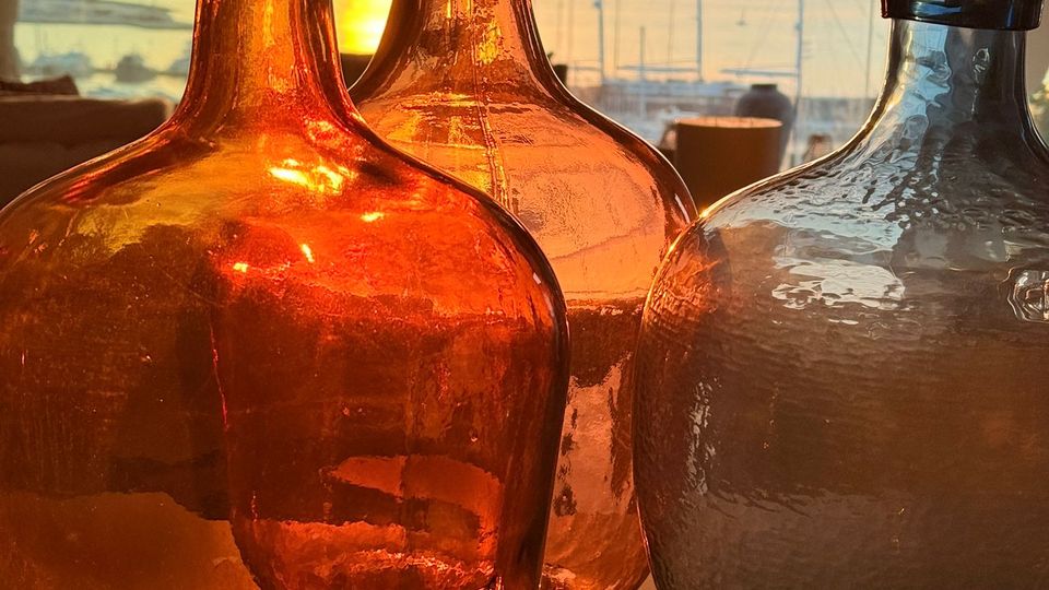 Große Glasflaschen in Bernstein- und Blautönen stehen im Vordergrund auf einem Tisch, dahinter ein Innenraum mit Hängeleuchten und großer Fensterfront. Durch die Fenster ist ein Sonnenuntergang über einem Hafen mit Segelmasten und Wasser zu sehen.