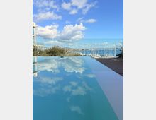 Infinity-Pool auf einer modernen Terrasse mit Glasgeländer, dessen Wasser die Wolken spiegelt. Im Hintergrund Blick über einen Yachthafen mit Segelmasten auf das Meer und eine entfernte Küstenstadt.