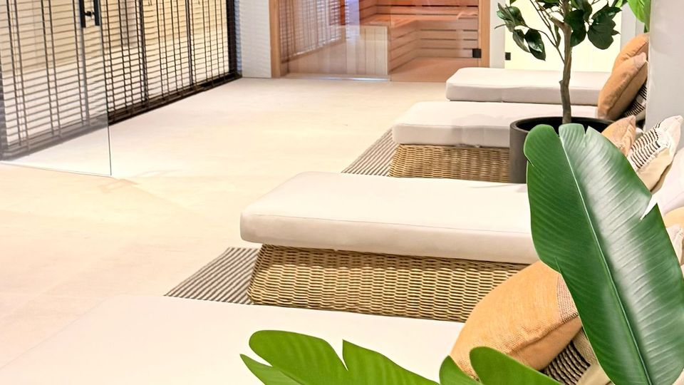 Moderner Wellness- und Ruhebereich mit weißen Liegen auf geflochtenen Rattan-Basen, hellen Fliesen und großen Grünpflanzen. Im Hintergrund sind durch Glaswände eine Sauna mit Holzverkleidung sowie ein angrenzender Bereich mit Lamellen- oder Gittertüren zu sehen.