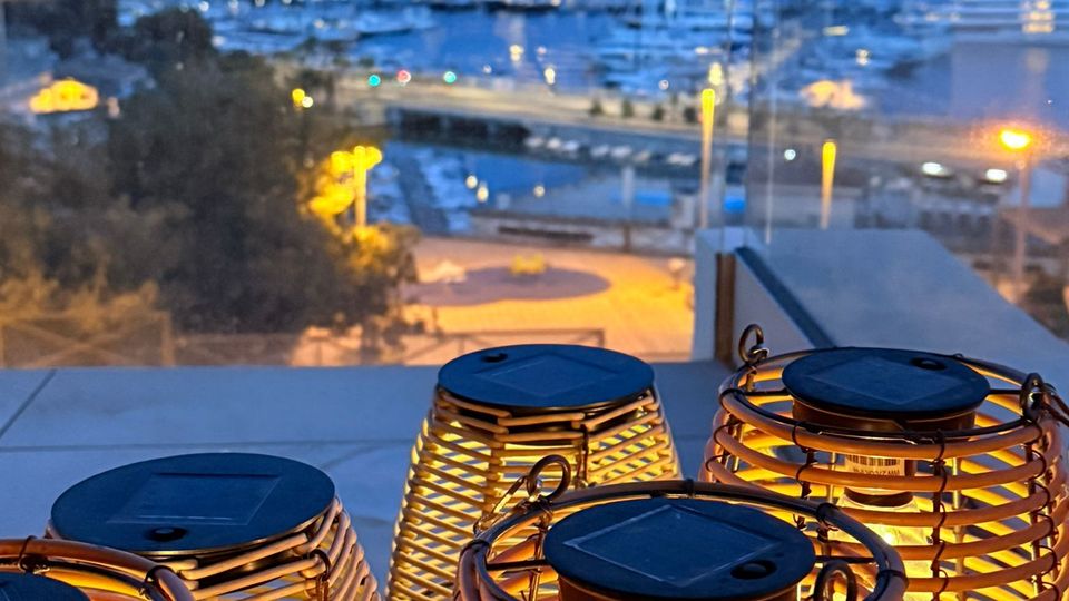 Blick von einer Terrasse oder einem Balkon auf einen beleuchteten Hafen mit Booten und Stadtlichtern in der Dämmerung. Im Vordergrund stehen mehrere leuchtende Laternen mit geflochtenen Holz- oder Rattan-Gittern auf einer Brüstung.