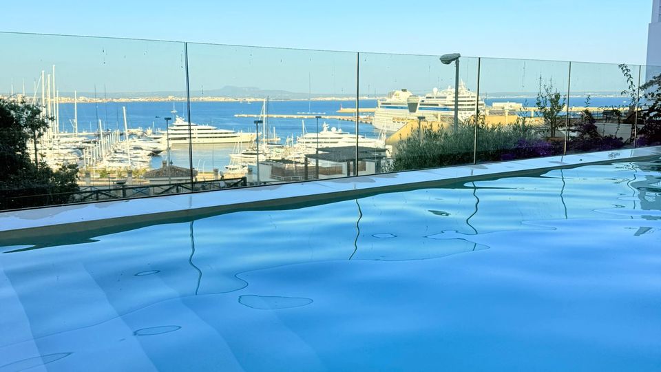 Infinity-Pool auf einer Terrasse mit Glasgeländer, im Vordergrund die ruhige Wasseroberfläche. Dahinter Blick auf einen Hafen mit zahlreichen Segelbootmasten, Yachten und einem großen Kreuzfahrtschiff vor Meer und Küstenlinie unter klarem Himmel.
