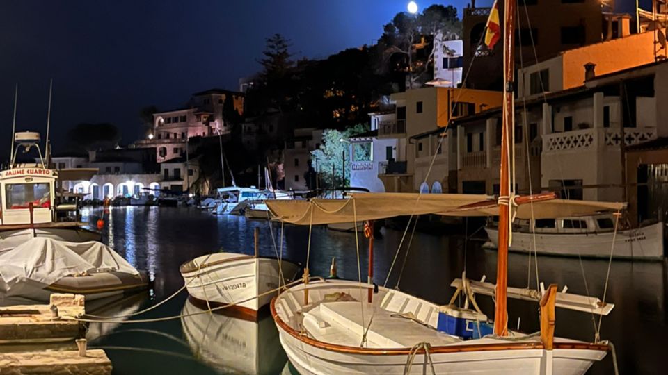 Port esportiu de nit amb petites barques blanques amarrades a l’aigua tranquil·la al costat d’un moll de pedra. Al fons hi ha edificis baixos il·luminats a la vora del canal i un turó amb arbres sota un cel fosc.
