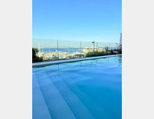 Infinity-Pool auf einer Terrasse mit Glasgeländer, im Vordergrund die ruhige Wasseroberfläche. Dahinter Blick auf einen Hafen mit zahlreichen Segelbootmasten, Yachten und einem großen Kreuzfahrtschiff vor Meer und Küstenlinie unter klarem Himmel.