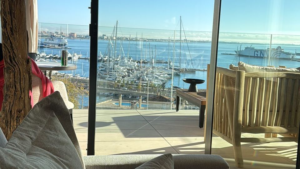 Modernes Wohnzimmer mit Sofa und Stehlampe vor bodentiefen Glasfronten, die auf eine Terrasse mit Holzdecke und Sitzmöbeln führen. Dahinter ist ein Yachthafen mit vielen Segelmasten und Blick auf das Meer unter blauem Himmel zu sehen.