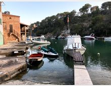 Petit port en una cala d’aigües tranquil·les, amb diverses embarcacions amarrades a un moll de pedra i una passarel·la estreta cap a un vaixell blanc. A l’esquerra hi ha edificis de pedra vora l’aigua i al fons un vessant boscós amb roques i pins que envolta la badia.