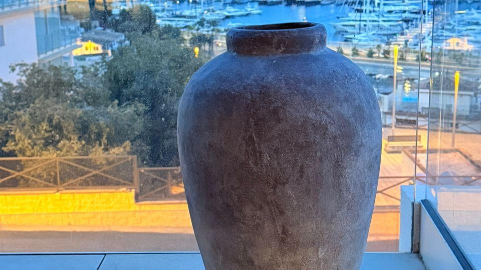 Blick von einer verglasten Terrasse auf einen beleuchteten Yachthafen mit vielen Masten und Booten in der Dämmerung. Im Vordergrund stehen eine große Tonvase auf Fliesen sowie kleine Laternen am Boden.