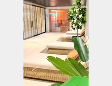 Moderner Wellness- und Ruhebereich mit weißen Liegen auf geflochtenen Rattan-Basen, hellen Fliesen und großen Grünpflanzen. Im Hintergrund sind durch Glaswände eine Sauna mit Holzverkleidung sowie ein angrenzender Bereich mit Lamellen- oder Gittertüren zu sehen.