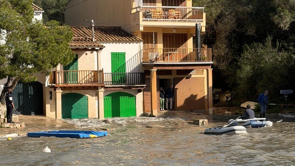 Edifici residencial de diverses plantes amb balcons i façana clara, amb portes verdes a la planta baixa, situat al costat d’un bosc. Davant de l’edifici hi ha una zona inundada amb aigua tèrbola i petites embarcacions flotant.