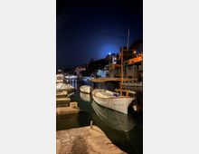 Port esportiu de nit amb petites barques blanques amarrades a l’aigua tranquil·la al costat d’un moll de pedra. Al fons hi ha edificis baixos il·luminats a la vora del canal i un turó amb arbres sota un cel fosc.