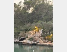 Escalinata i camí de pedra que baixen fins a una petita cala d’aigua tranquil·la, amb murs de contenció de roca i fanals encesos. Al fons hi ha un vessant boscós dens amb arbres i matolls sobre les roques de la costa.