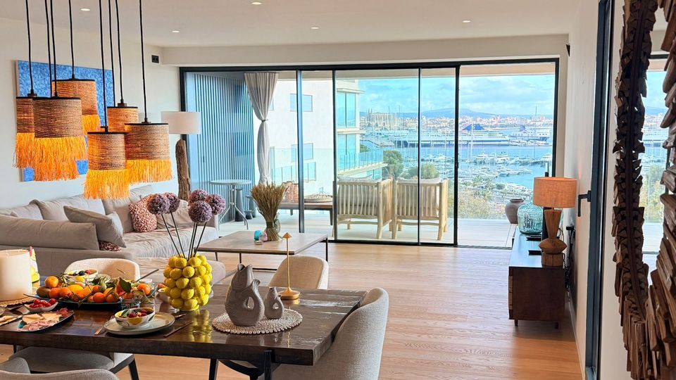 Offener Wohn- und Essbereich in einer modernen Wohnung mit hellem Holzboden, Esstisch mit Polsterstühlen und Sofaecke unter mehreren hängenden Lampenschirmen. Große Schiebetüren führen auf einen Balkon; durch die Glasfront ist ein Blick auf eine Marina und das Wasser zu sehen.